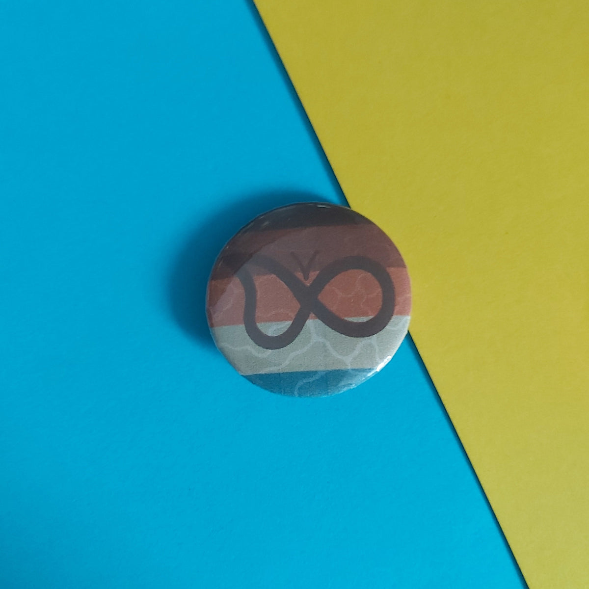 A round pin badge featuring a stylized infinity symbol over a horizontal tricolor flag of brown, orange, and light blue, placed on a diagonally split blue and yellow background.