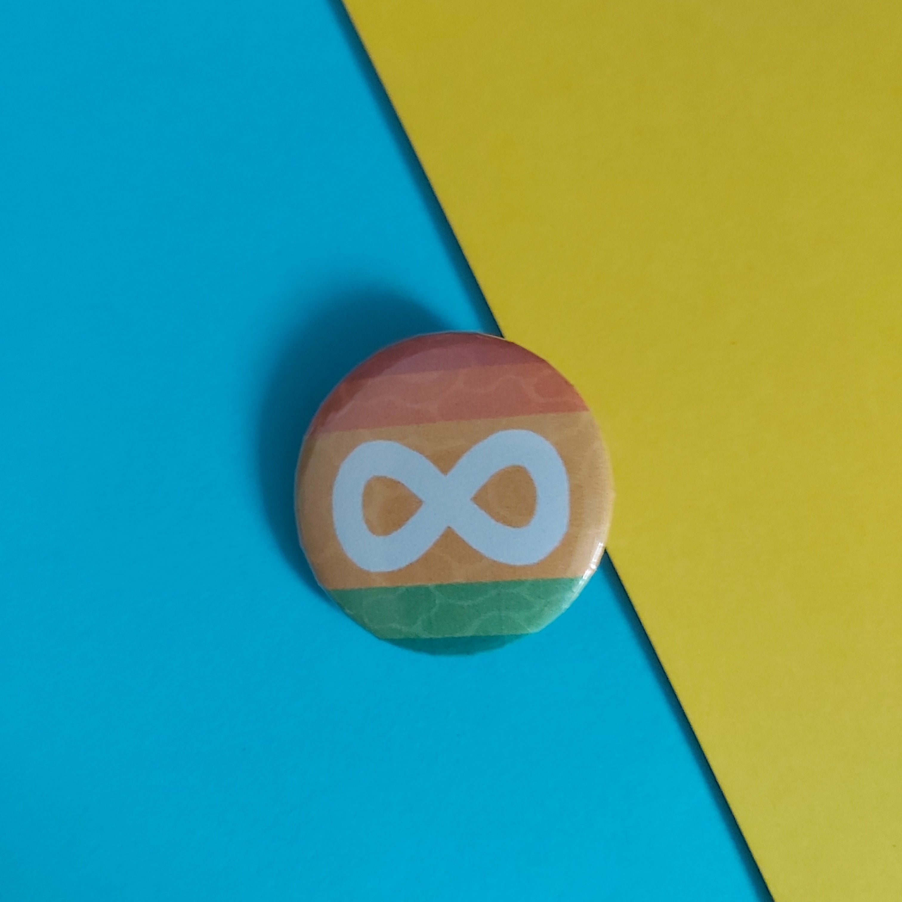 A small round pinback button featuring the white infinity symbol, set against a diagonally split bright blue and yellow background.