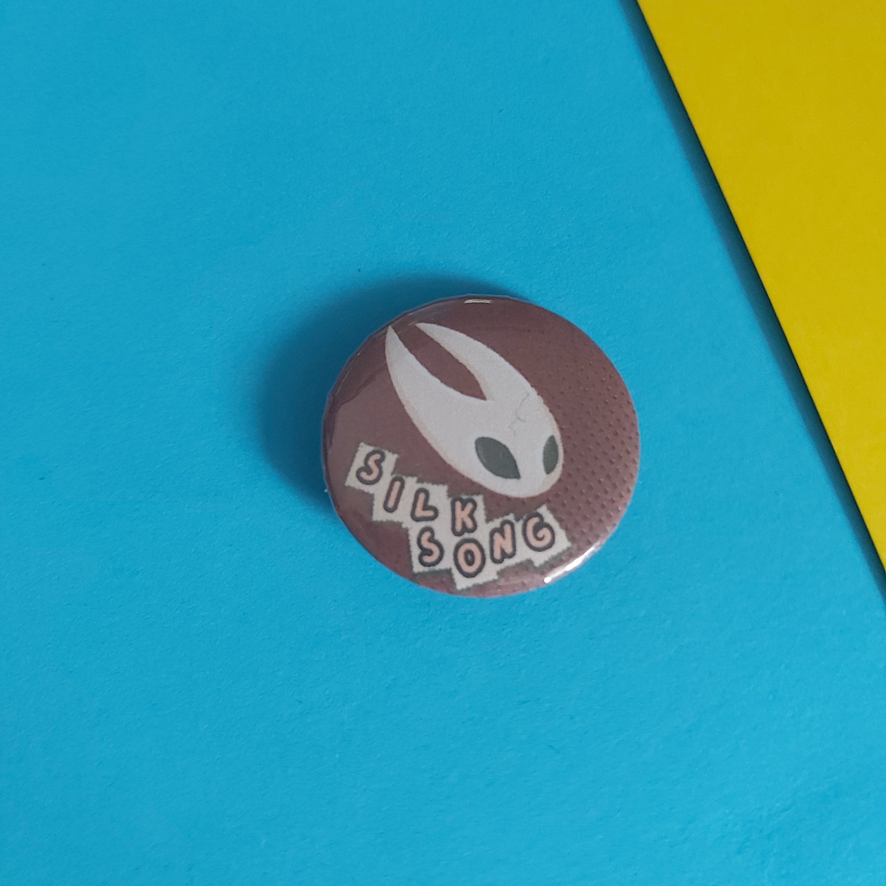 A round pinback button featuring the 'Silk Song' artwork, displayed on a bright blue background with a yellow corner accent.