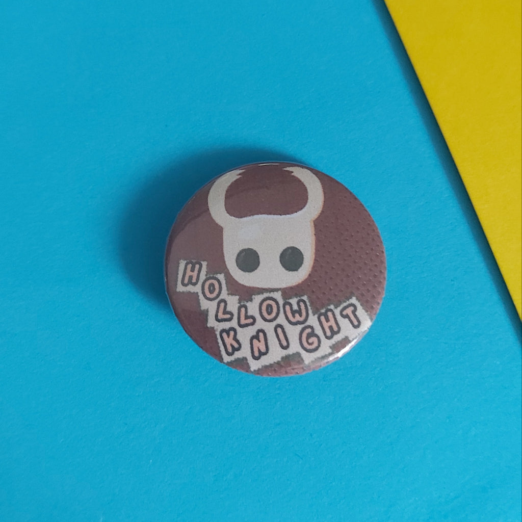 A round pinback button badge featuring a white skull with horns and the text 'HOLLOW KNIGHT' on a dark brown background, placed on a bright blue surface with a yellow corner visible in the upper right.
