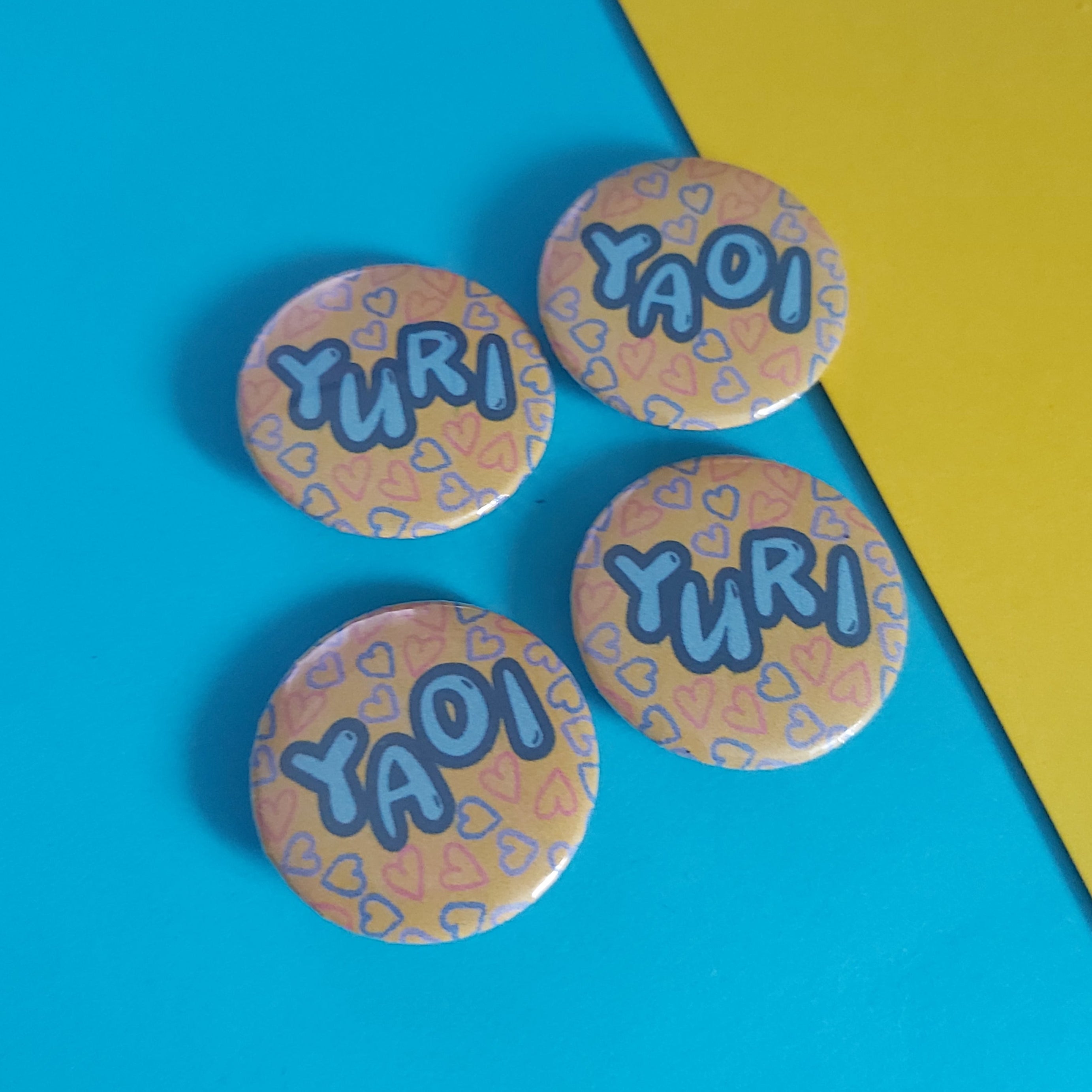 Four round pin-back buttons featuring the words 'YURI' and 'YAOI' in blue bubble letters over a peach and blue patterned background, arranged on a split blue and yellow background.