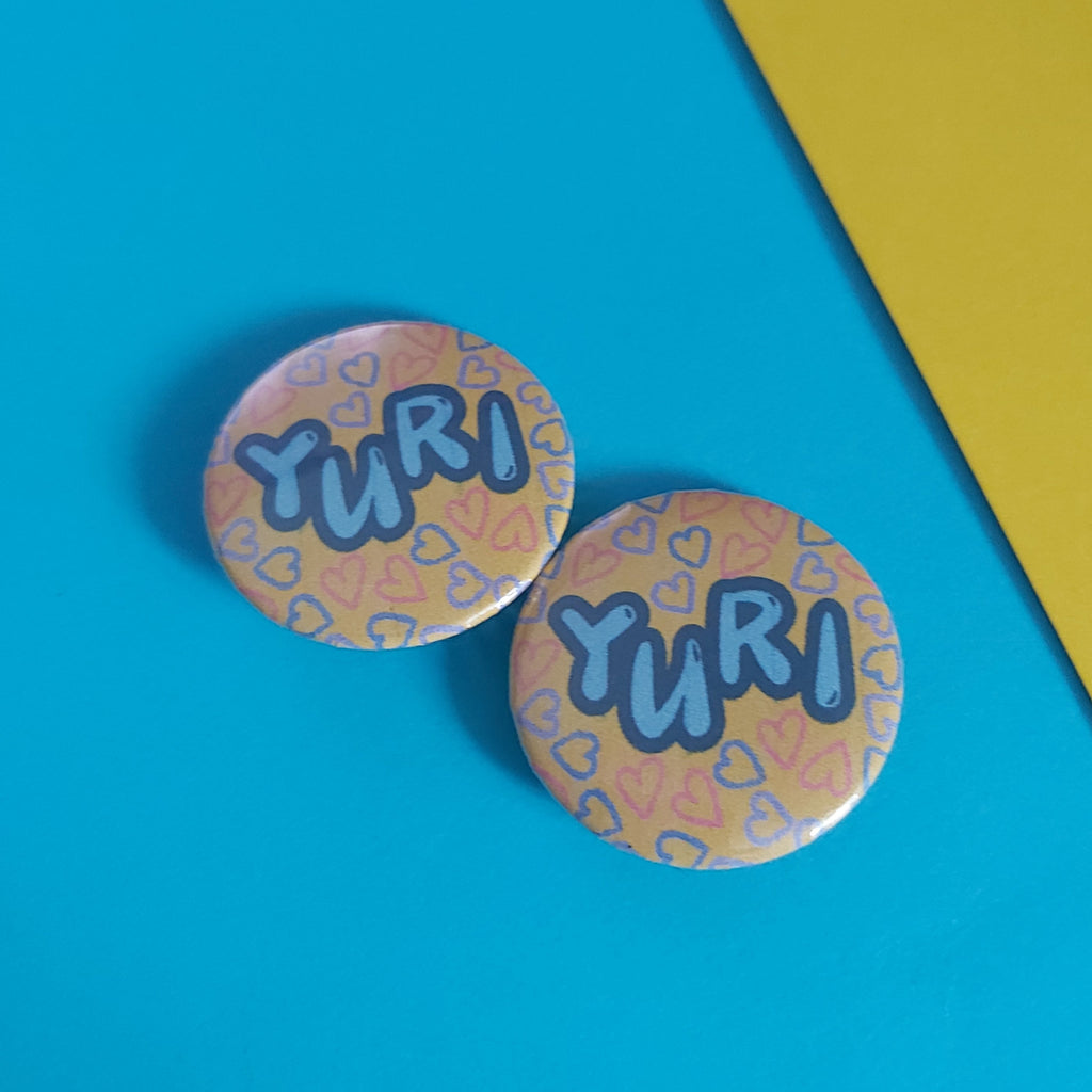 Two round pin-back buttons with the name 'YURI' in blue bubble letters over a pink and orange heart patterned background, displayed on a bright blue surface with a yellow corner visible.