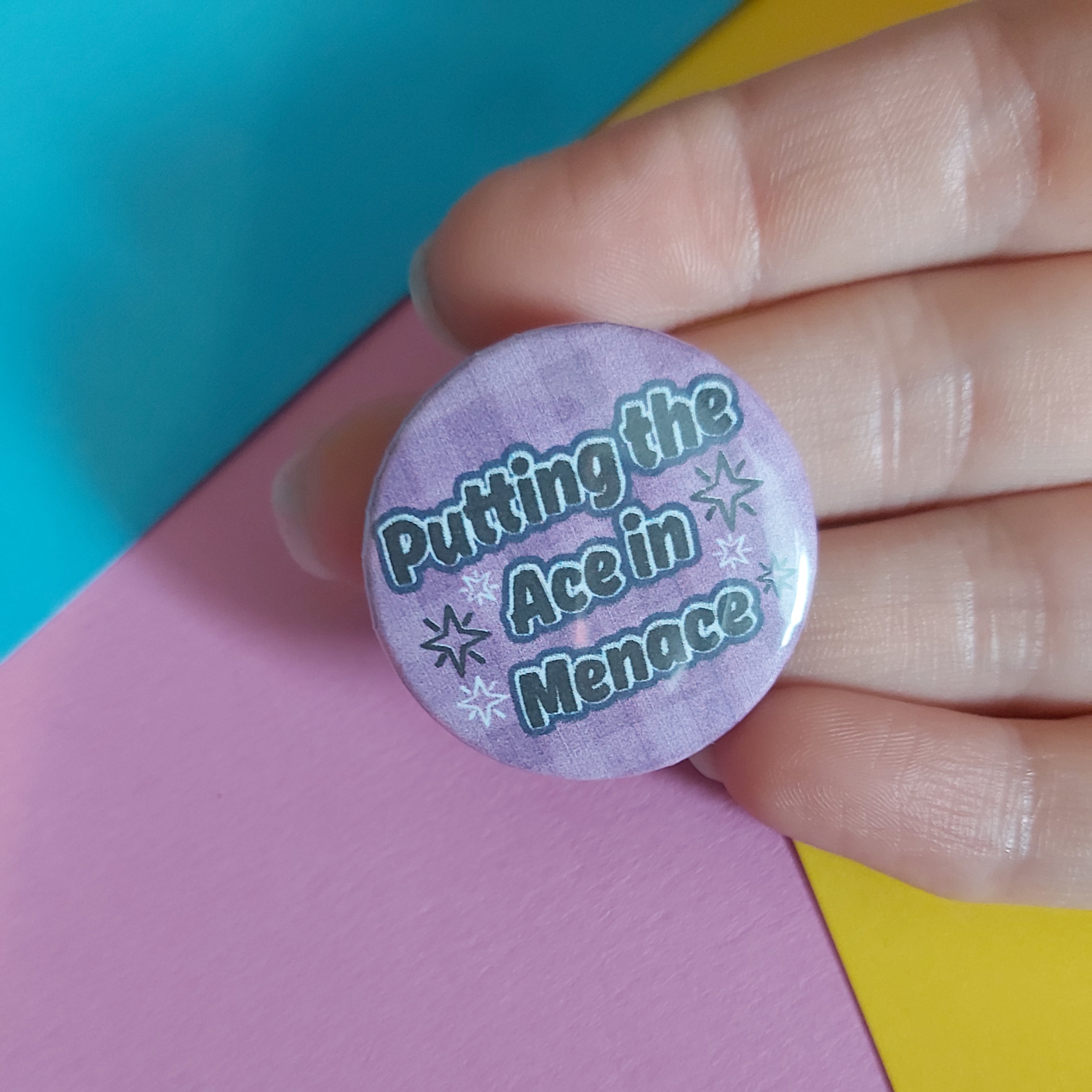 A hand holding a round pinback button badge with a lavender background and the text 'Putting the Ace in Menace' in white with a teal outline, set against a background of pink, yellow, and teal paper.