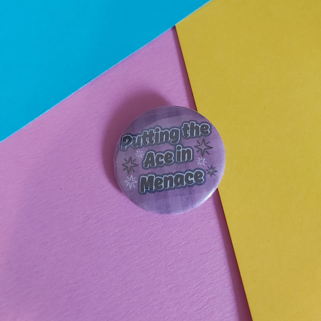 A round pinback button badge with the text 'Putting the Ace in Menace' in dark lettering on a light purple background, styled on a background of three colored paper sheets: cyan, pink, and yellow.