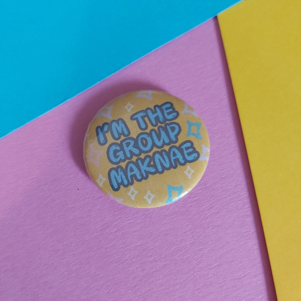 A round pin badge with the text 'I'M THE GROUP MAKNAE' in blue letters on a yellow background with light blue pattern details, resting on a diagonally split pink and turquoise background with a yellow corner.