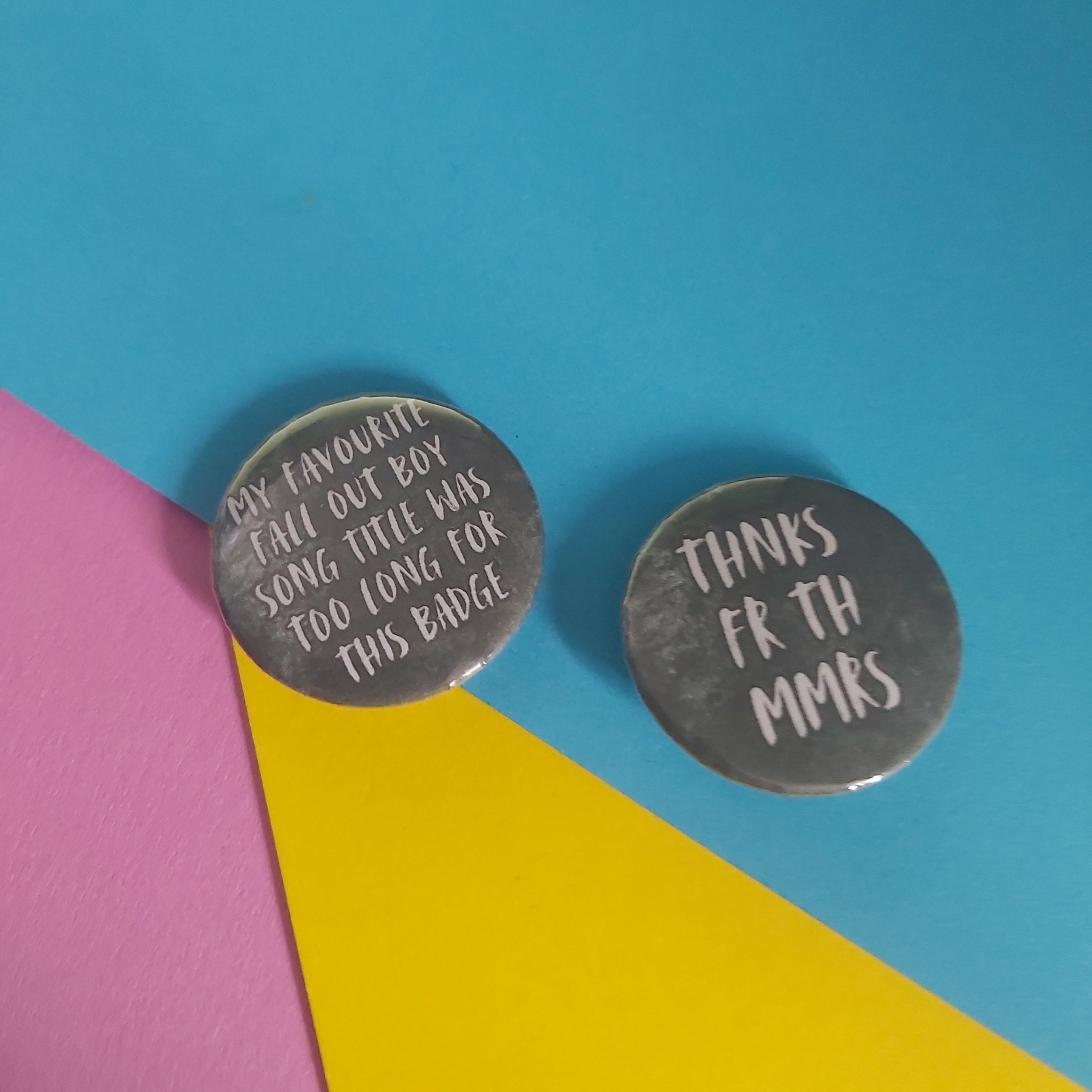 Two round emo-themed badges with text, displayed on a colourful background split diagonally into pink, yellow, and blue sections.