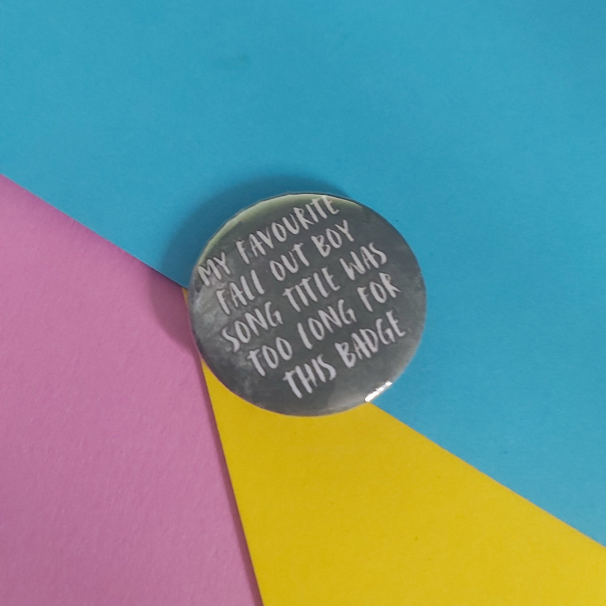 A round pin badge with white handwritten text reading 'MY FAVOURITE FALL OUT BOY SONG TITLE WAS TOO LONG FOR THIS BADGE', placed on a colorful background of cyan, pink, and yellow paper.