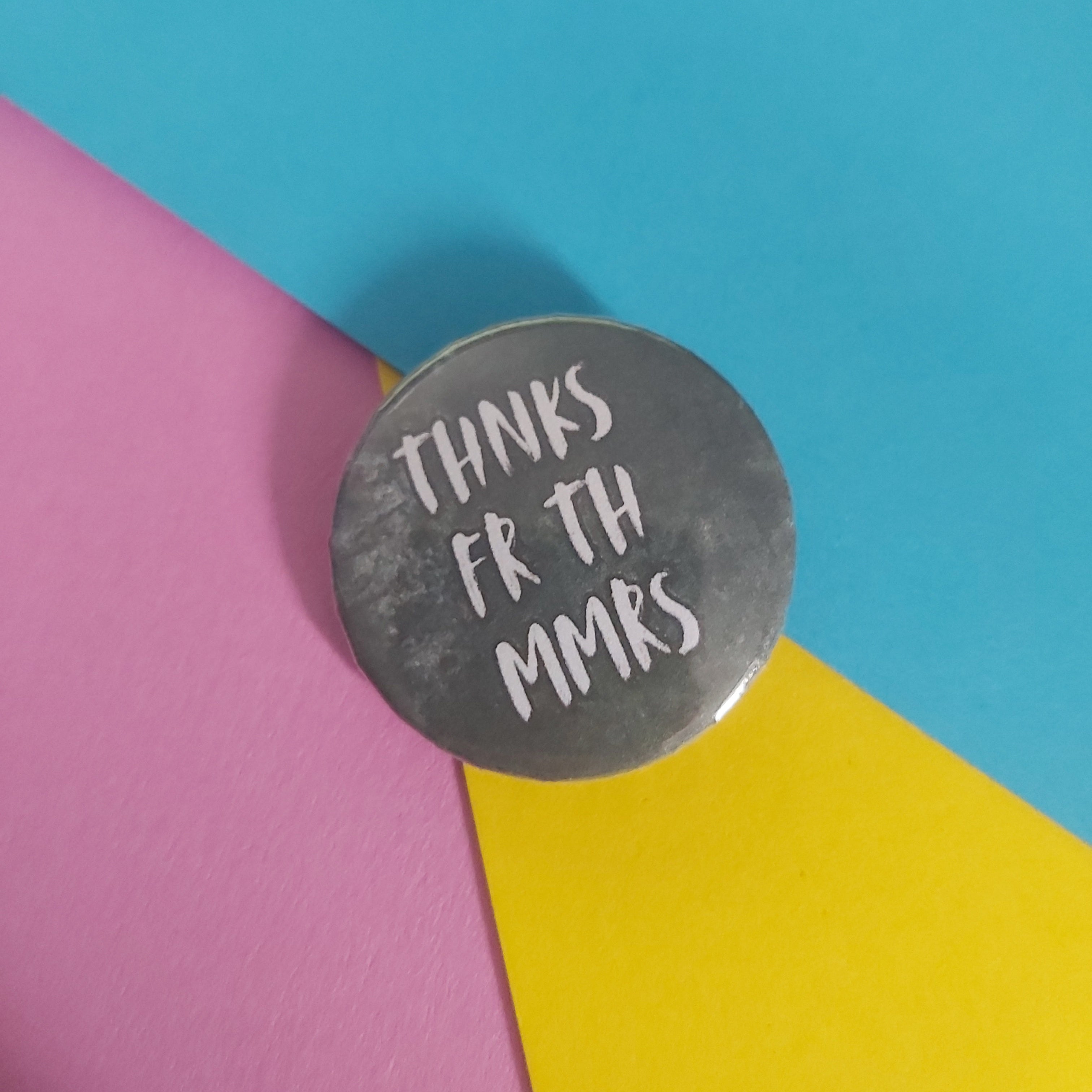 A round grey pin with the text 'THANKS FR TH MMRS' in white lettering, placed on a background of pink, blue, and yellow geometric paper.