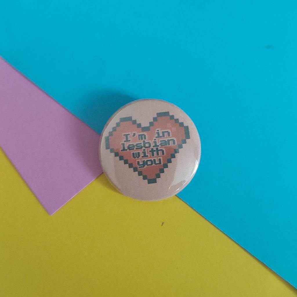 A round pinback button badge featuring a pixelated pink heart with the text 'I'm in lesbian with you' in black, placed on a background of bright yellow, cyan, and pink paper.