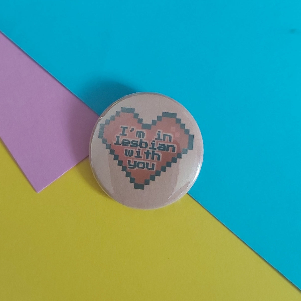 A round pin badge with a pixelated pink heart and the text 'I'm in Lesbian with you' on a light peach background, styled on a colourful background of cyan, yellow, and pink paper.