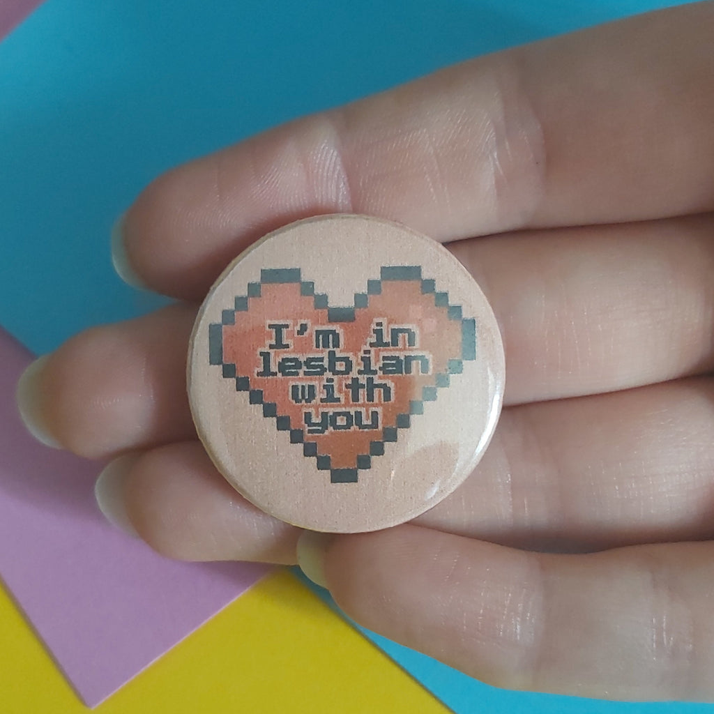 A hand holding a round pinback button with a pixelated heart and the text 'I'm in lesbian with you' in pink, set against a background of colourful paper sheets.
