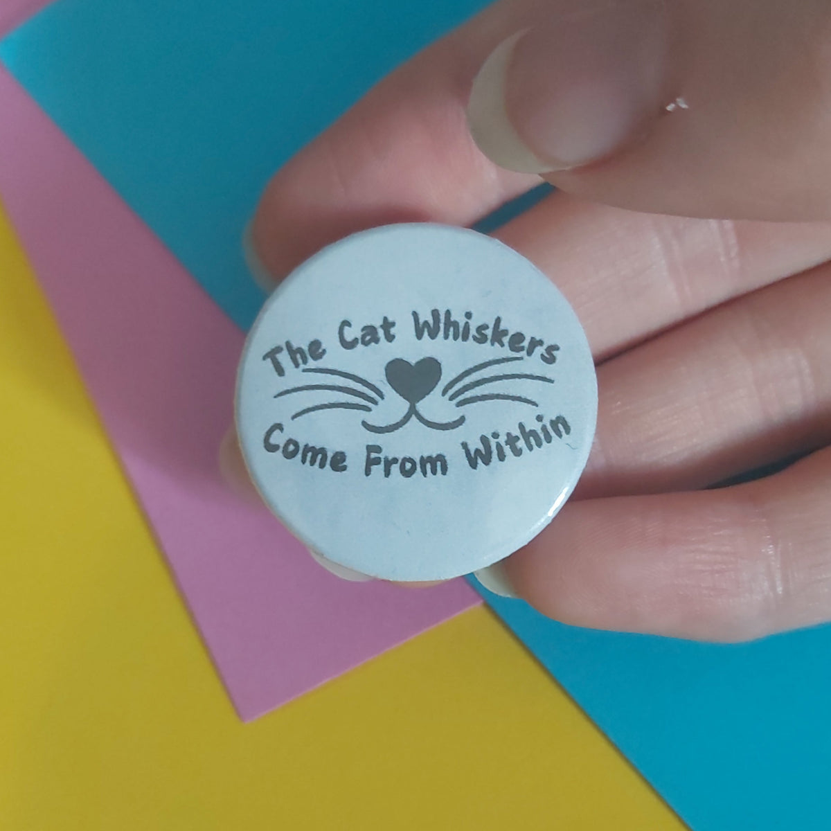 A hand holding a round white pin badge with black text that reads 'The Cat Whiskers Come From Within' and a simple whisker-and-heart nose graphic, set against a colourful background of yellow, pink, and blue paper.