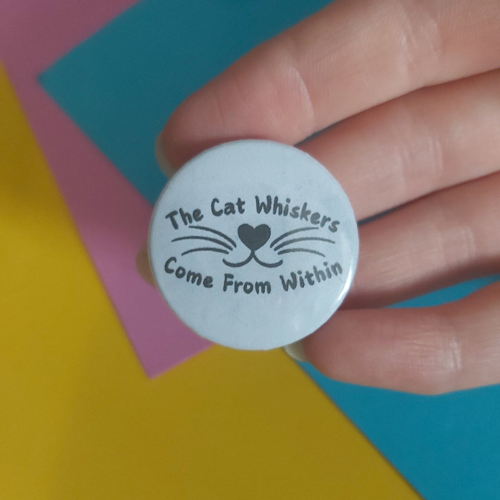 A hand holding a round white button or sticker with black text reading 'The Cat Whiskers Come From Within' and a simple whisker-and-heart graphic, against a colorful background of yellow, pink, and blue sheets.