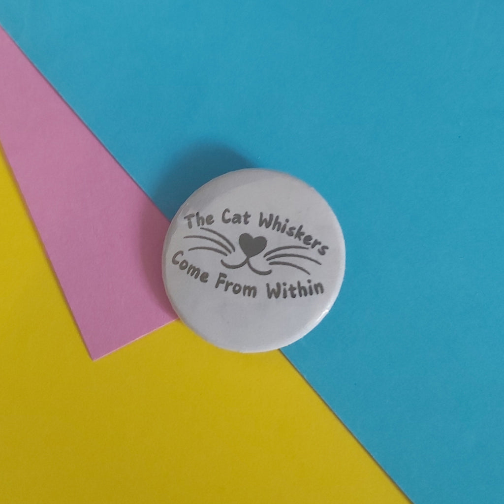 A round white pinback button with black text reading 'The Cat Whiskers Come From Within' and a simple whisker graphic, placed on a colorful background of pink, yellow, and blue paper.