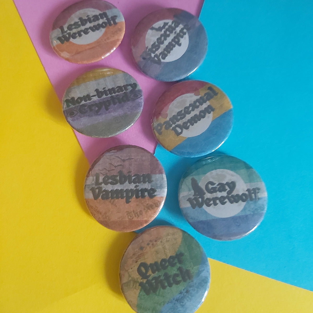 Seven round pinback buttons with various LGBTQ+ themed phrases like 'Lesbian Werewolf', 'Non-binary cryptid', and 'Queer Witch' arranged on a bright yellow and blue background.