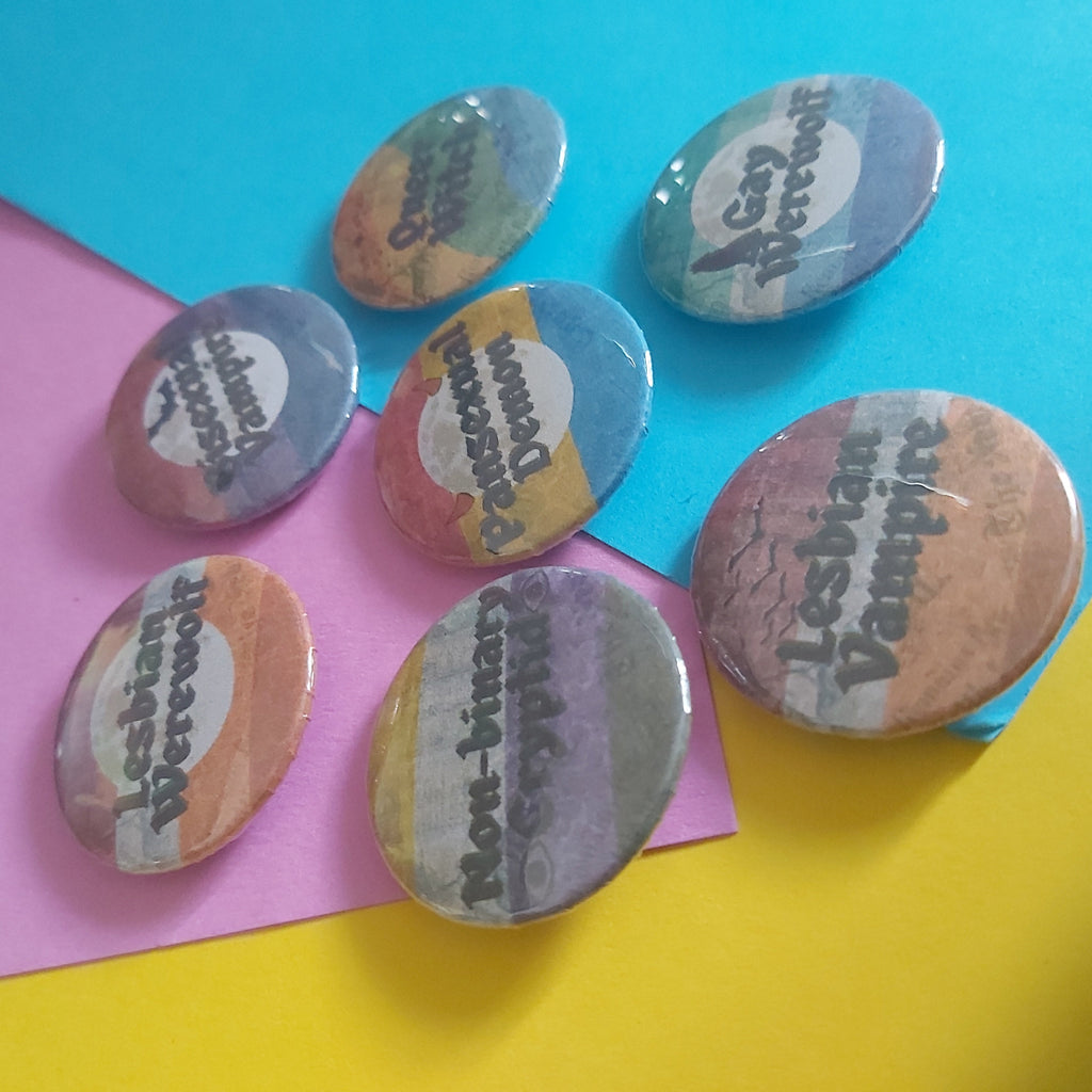 Six round button badges with printed pride-themed supernatural being phrases are arranged on bright colored paper backgrounds.
