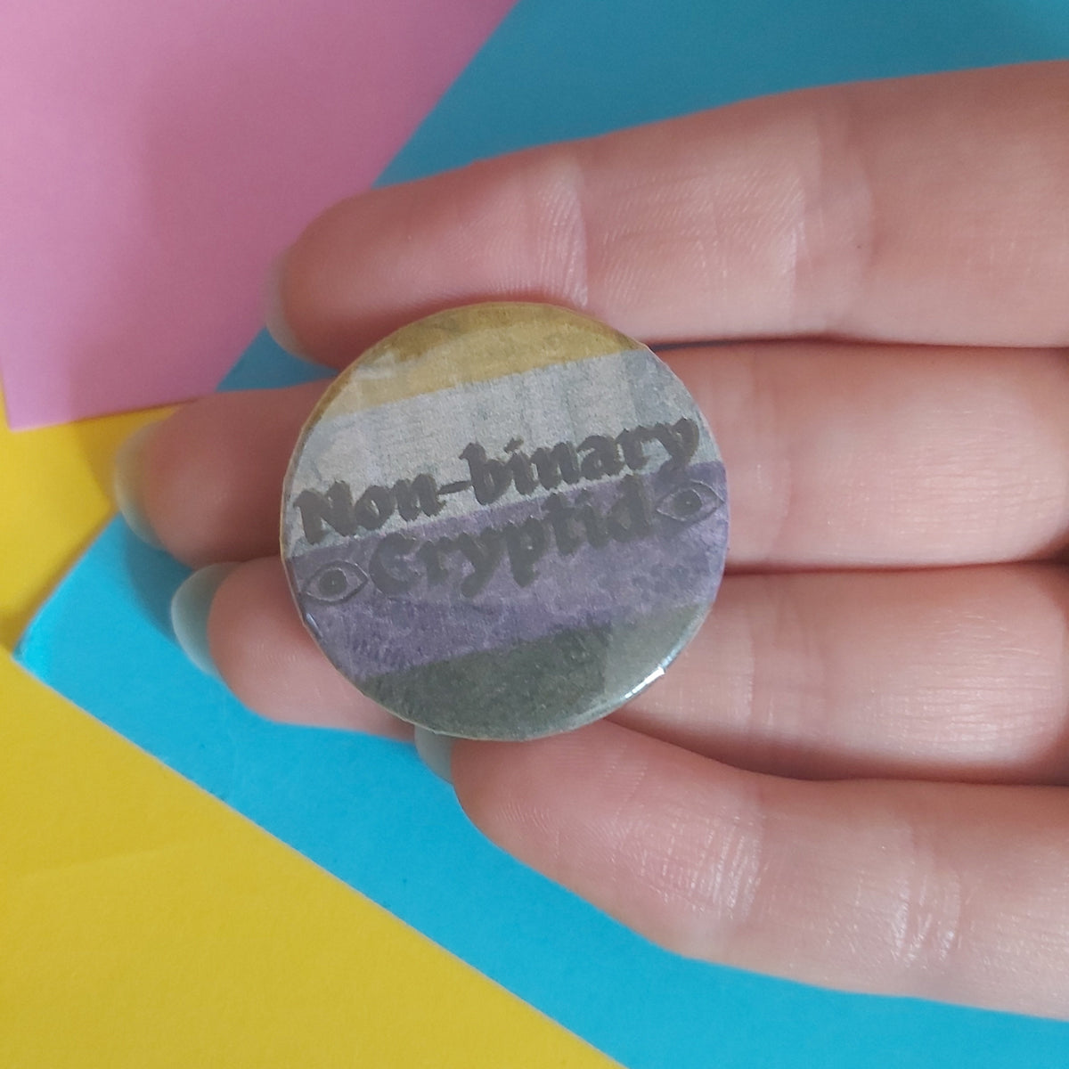 A hand holding a round pinback button with a non-binary pride flag design and the text 'Non-binary Crypid'. The background consists of colourful paper sheets in pink, blue, and yellow.