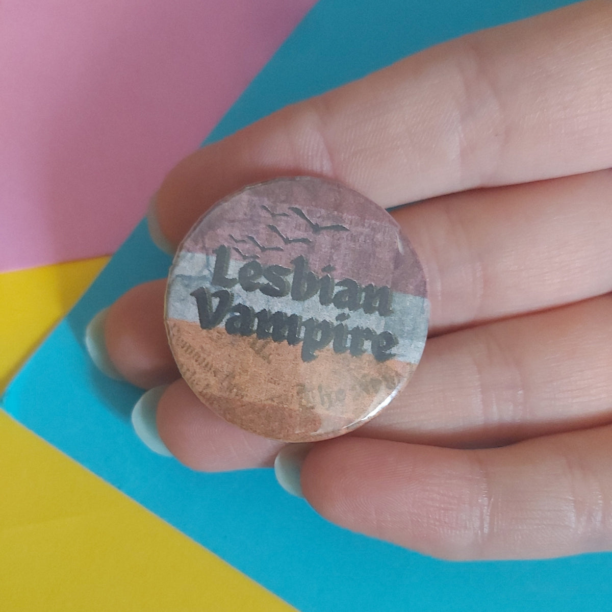 A hand holding a round pinback button badge with a distressed design featuring the words 'Lesbian Vampire' in dark gray text