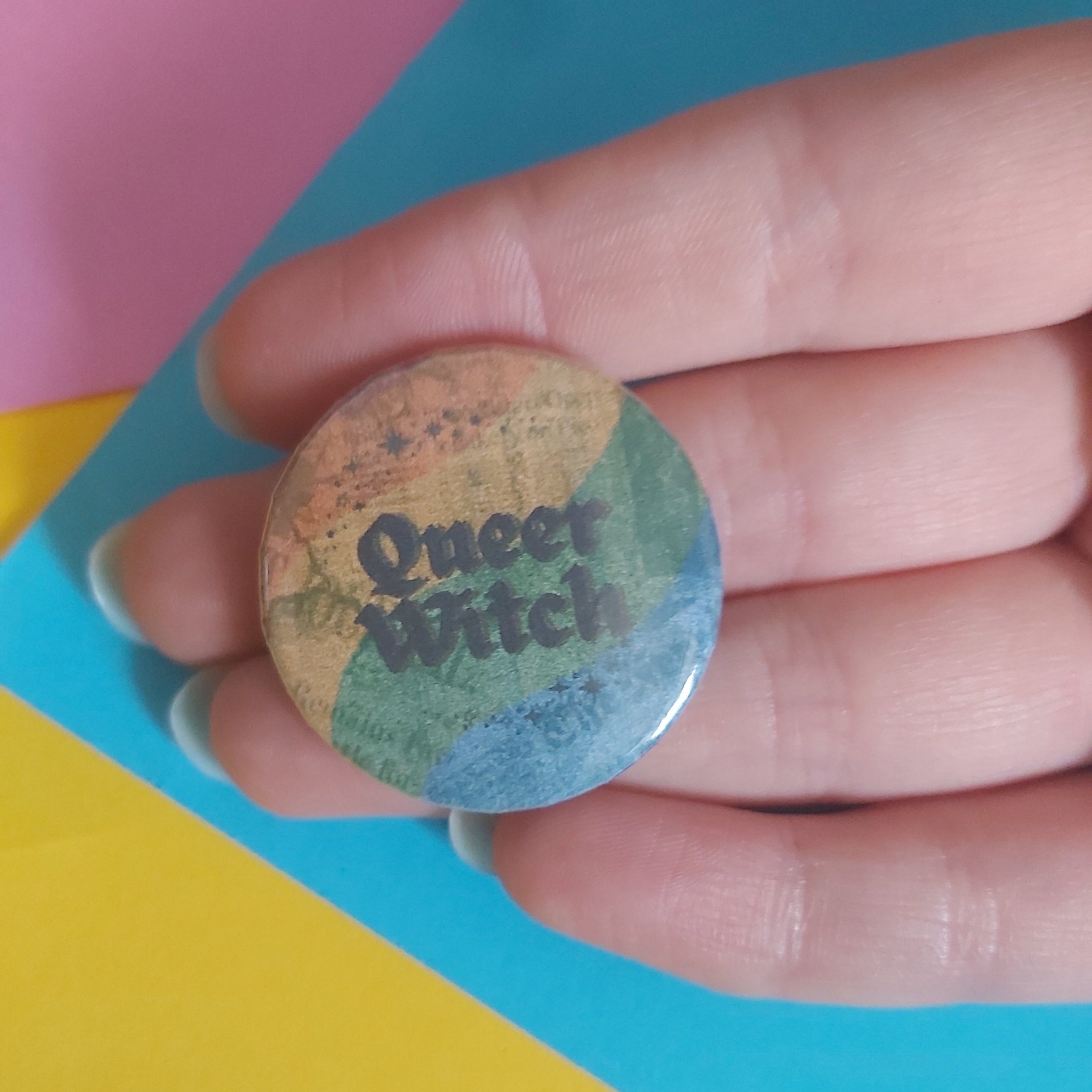 A hand holding a round pinback button badge with a muted rainbow background and the words 'Queer Witch' in dark lettering, set against a colorful background of pink, blue, and yellow paper.