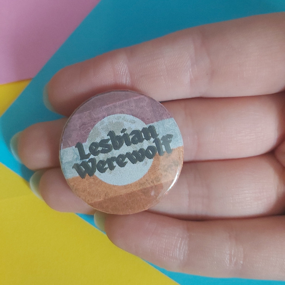 A hand holding a round pinback button badge with a pink, white, and orange design and the text 'Lesbian Werewolf' in black script, set against a background of pink, blue, and yellow paper.