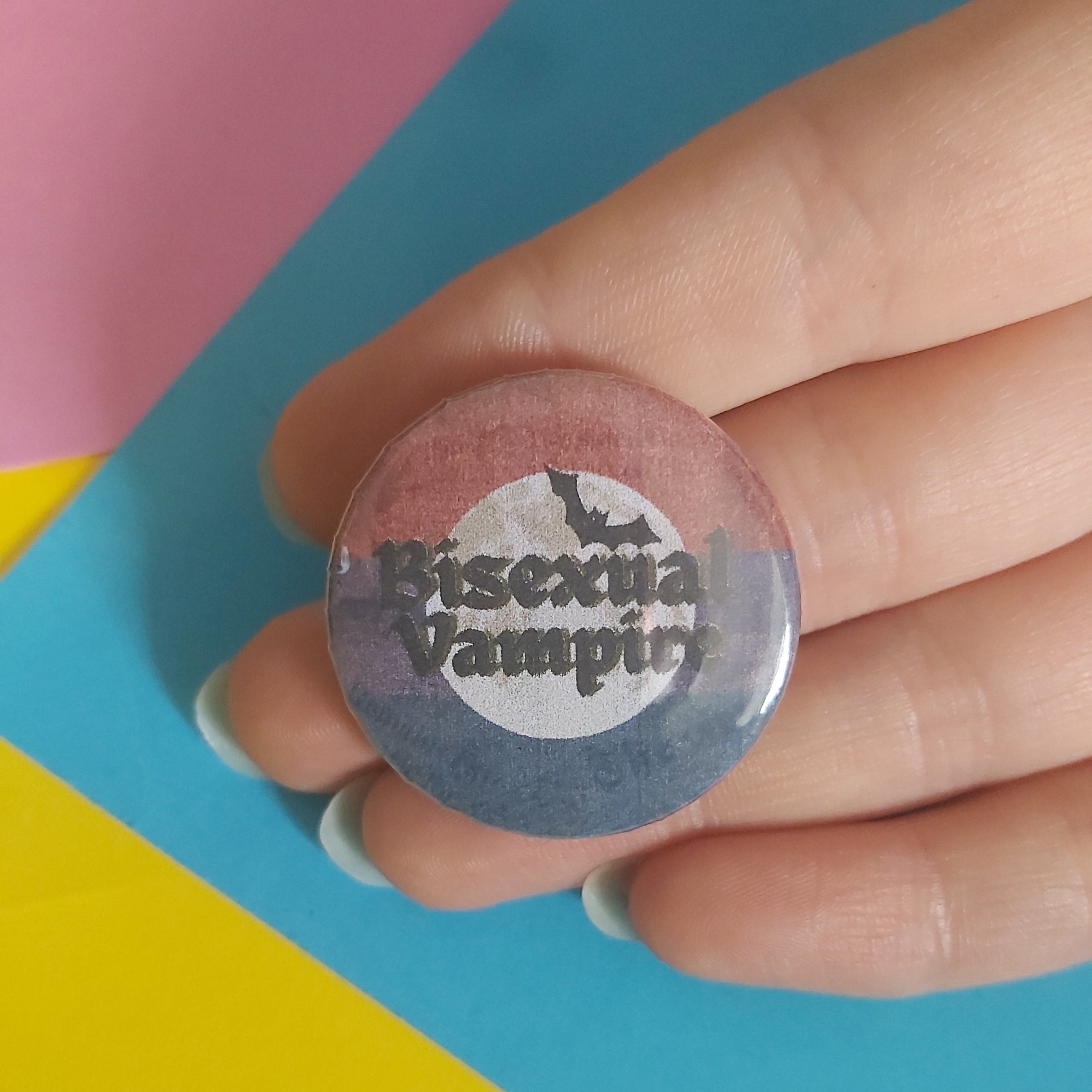 A hand holding a round pinback button badge with the text 'Bisexual Vampire' and a bat graphic, set against a colorful background of pink, blue, and yellow paper.