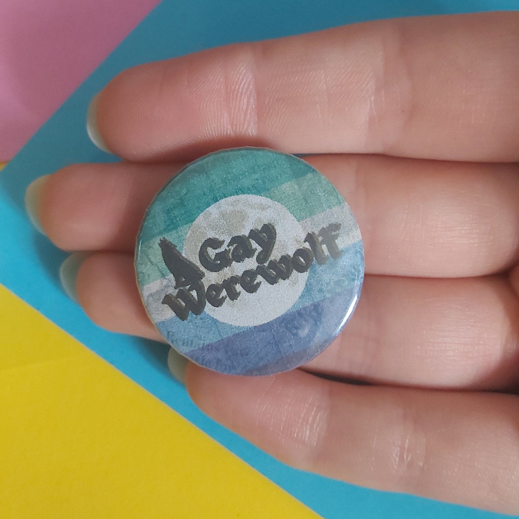 A small round pinback button held in a hand, featuring a design with teal, blue, and white tones and the text 'Gay Werewolf' in black script.