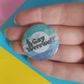 A small round pinback button held in a hand, featuring a design with teal, blue, and white tones and the text 'Gay Werewolf' in black script.