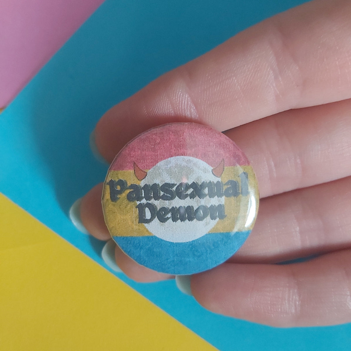 A hand holding a round pinback button featuring the Pansexual Flag colours with the text 'Pansexual Demon' in black on a full moon motif.