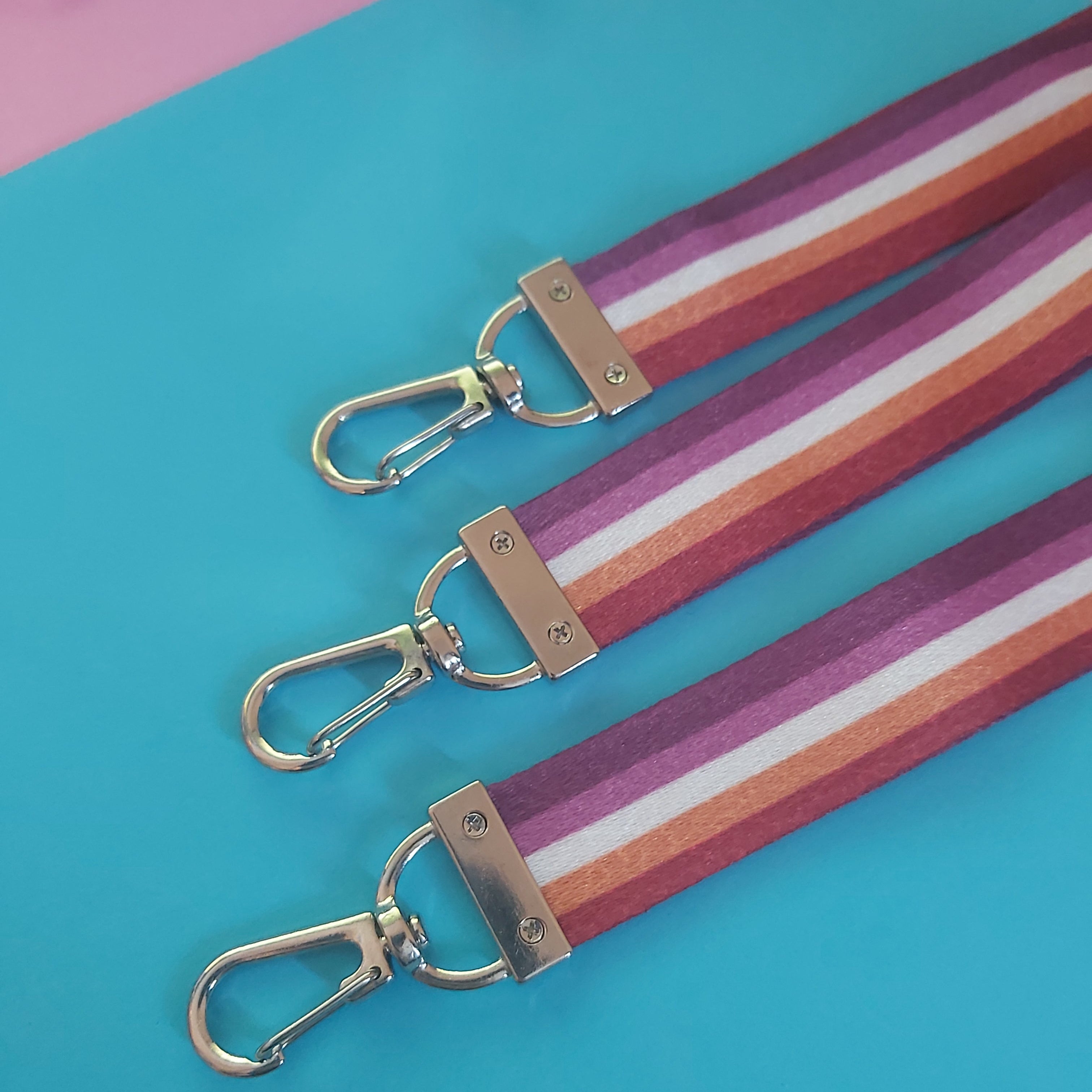 Three fabric keychains with silver-tone swivel clasp hardware, featuring diagonal stripes of orange, pink, and white, arranged on a bright blue background with a pink corner.