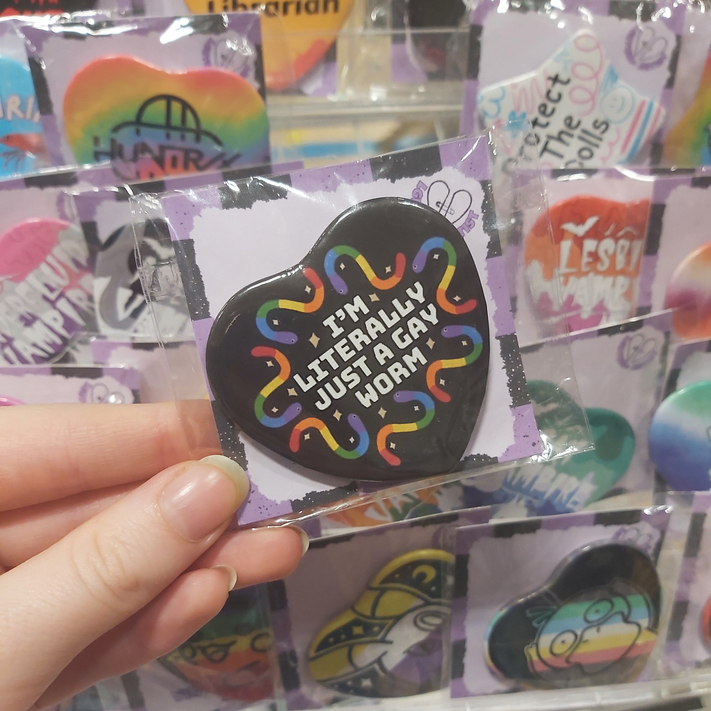 A hand holding a heart-shaped black pin with colourful squiggly worms and the text 'I'M LITERALLY JUST A GAY WORM' in front of a display of other pins.