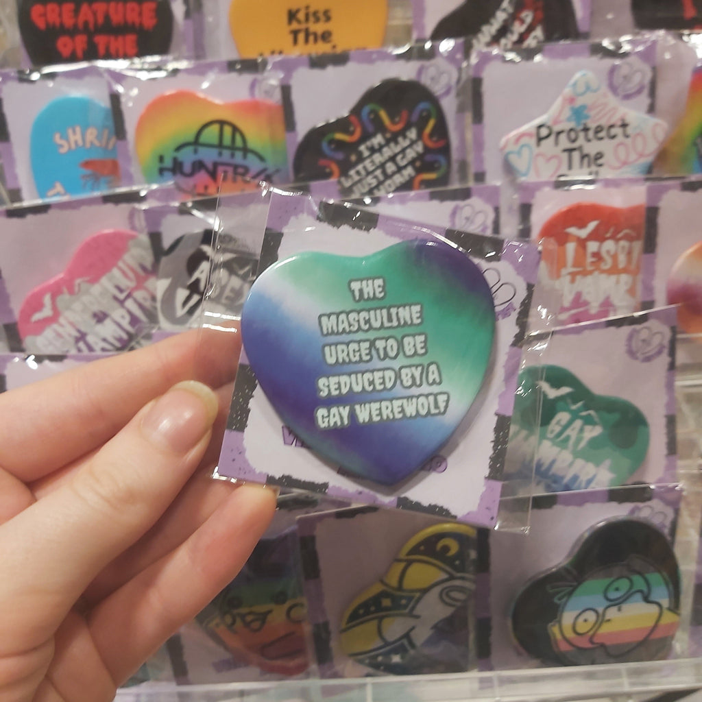 A hand holding a heart-shaped pin with a blue-to-green gradient background and the text 'THE MASCULINE URGE TO BE SEDUCED BY A GAY WEREWOLF'. In the background, a retail display rack shows other colourful shaped pins.