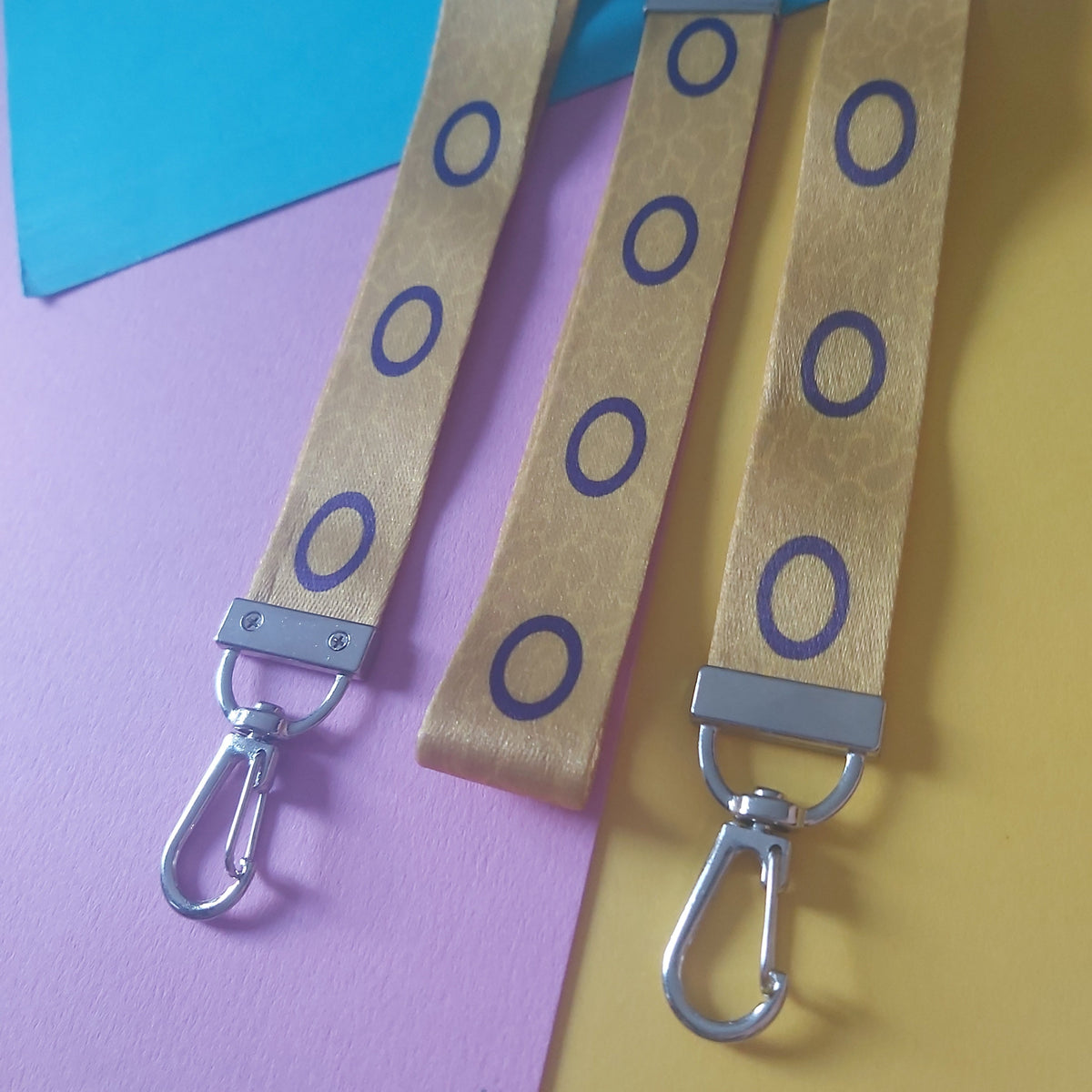 Three yellow cotton wristlets with purple circular patterns and silver metal carabiners, arranged diagonally on a split pink and yellow background with a blue corner accent.