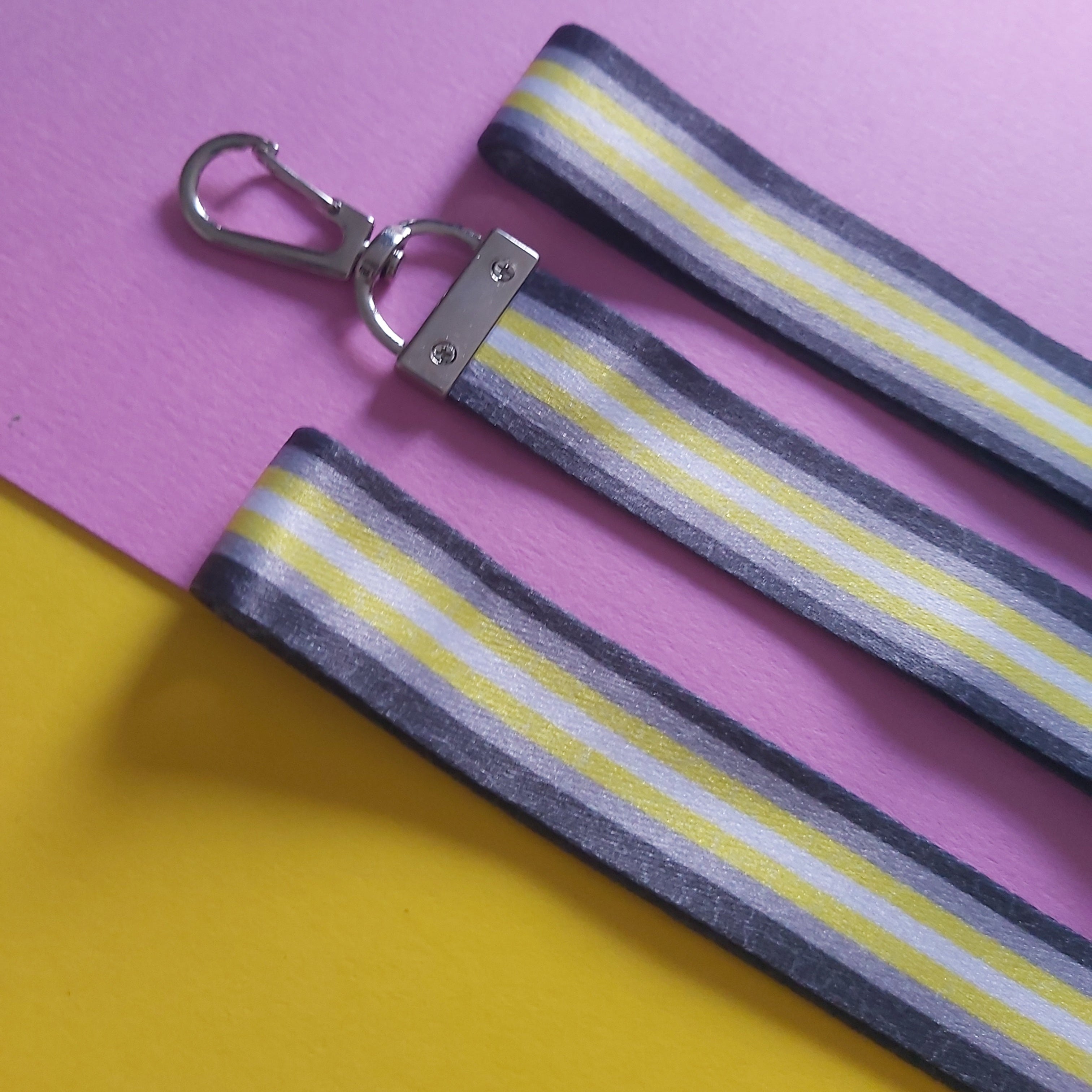 Three fabric strap accessories with a grey, white, and yellow striped pattern and silver metal clasp, laid flat on a split pink and yellow background.