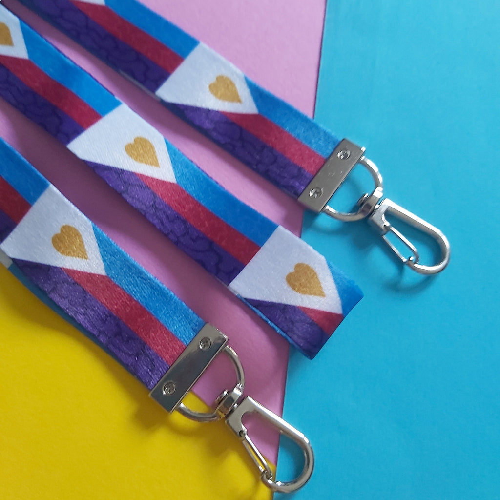 Three fabric keychains with silver metal clasp hooks, featuring a colourful pattern of blue, pink, and purple stripes with white triangles and gold hearts, laid out on a split pink, yellow, and blue background.