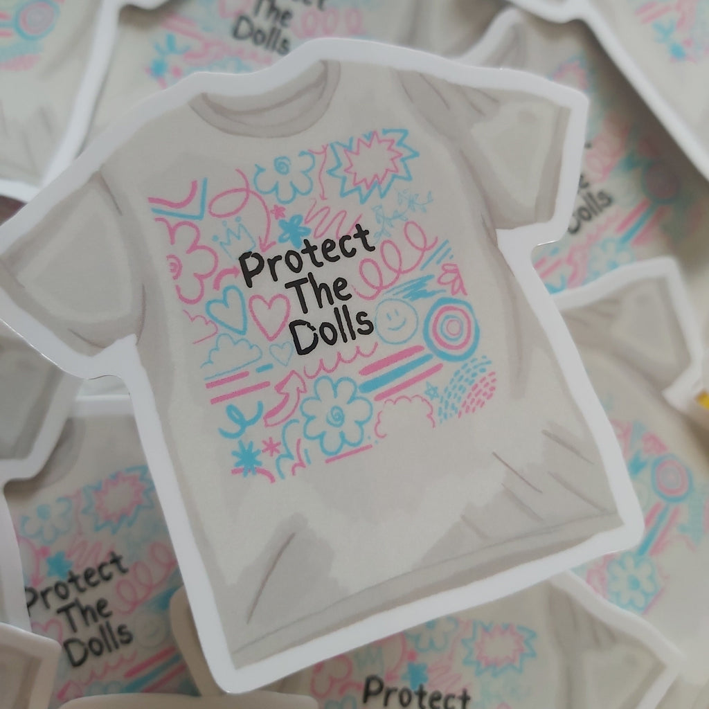 A die-cut sticker shaped like a t-shirt, featuring the text 'Protect The Dolls' in black alongside pink and blue doodle graphics, displayed on top of other matching stickers.