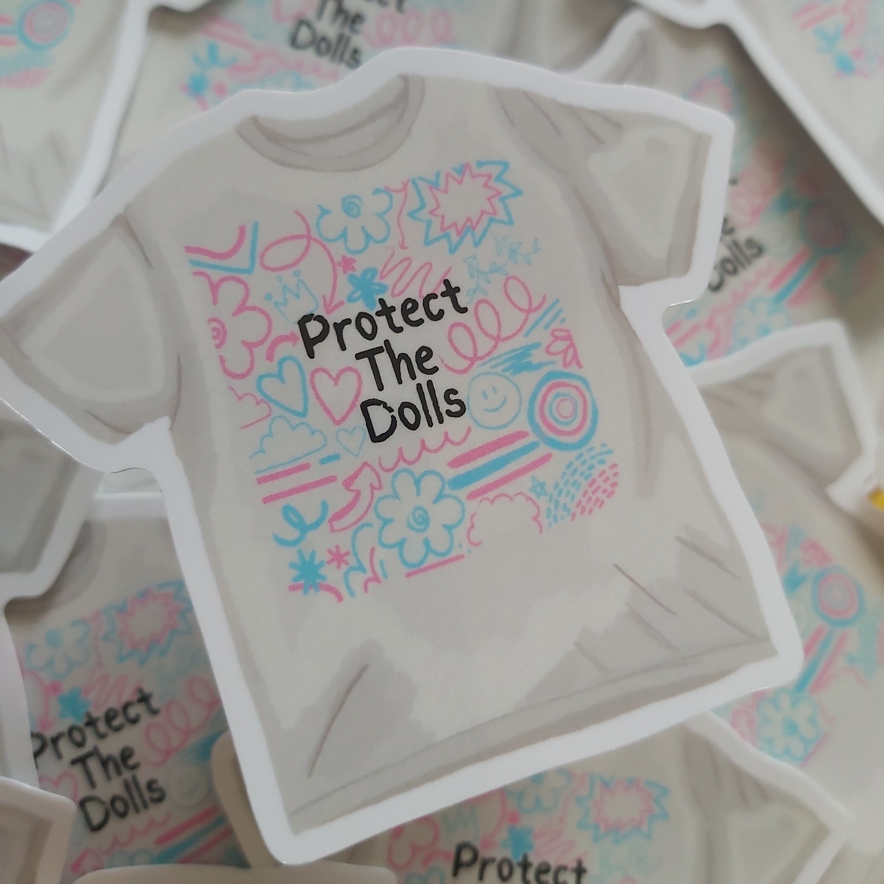 A die-cut sticker shaped like a t-shirt, featuring the text 'Protect The Dolls' in black alongside pink and blue doodle graphics, displayed on top of other matching stickers.