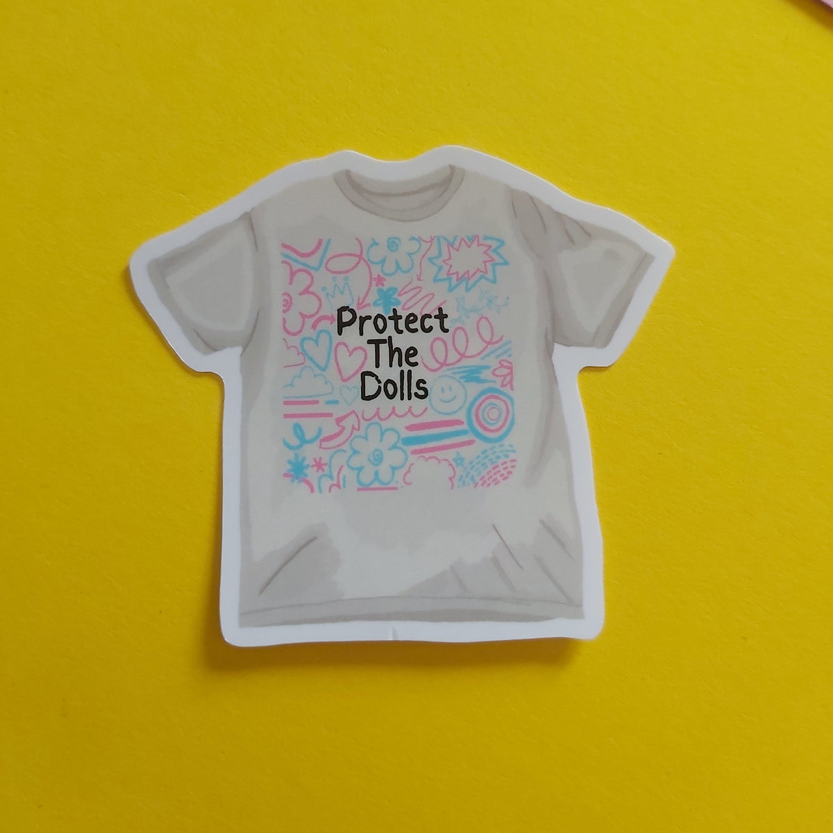 A die-cut sticker shaped like a white t-shirt, featuring the text 'Protect The Dolls' in black over a colourful pink and blue doodle pattern. The sticker is displayed on a bright yellow background with a pink corner visible in the top right.