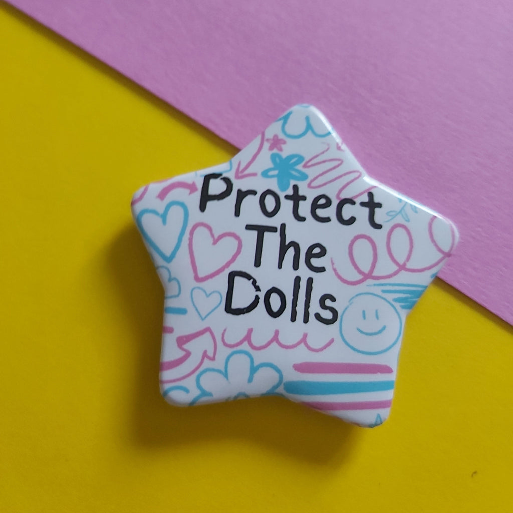 A star-shaped enamel pin with the text 'Protect The Dolls' in black, decorated with pink and blue doodles on a white background, set against a split yellow and pink backdrop.