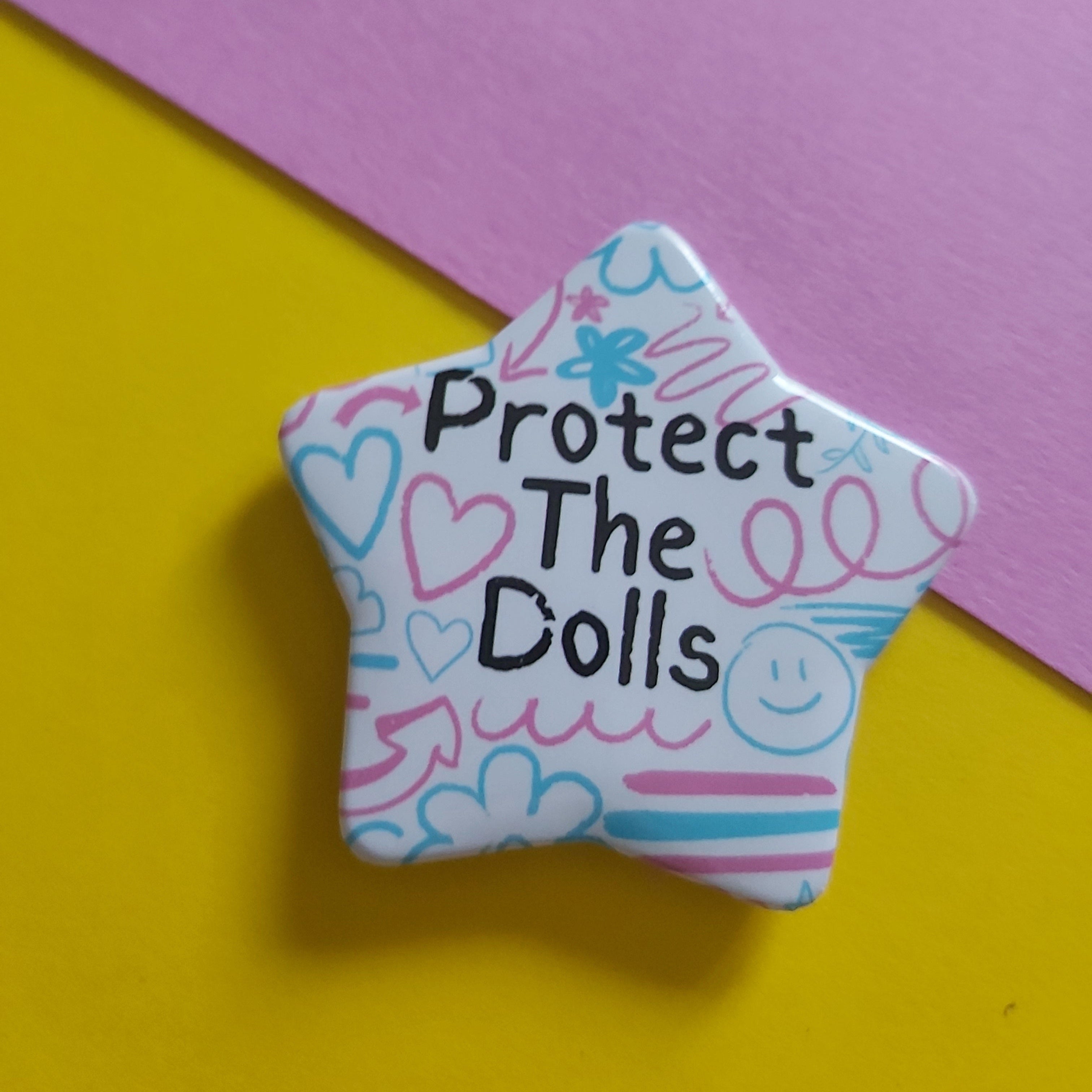 A star-shaped enamel pin with the text 'Protect The Dolls' in black, decorated with pink and blue doodles on a white background, set against a split yellow and pink backdrop.