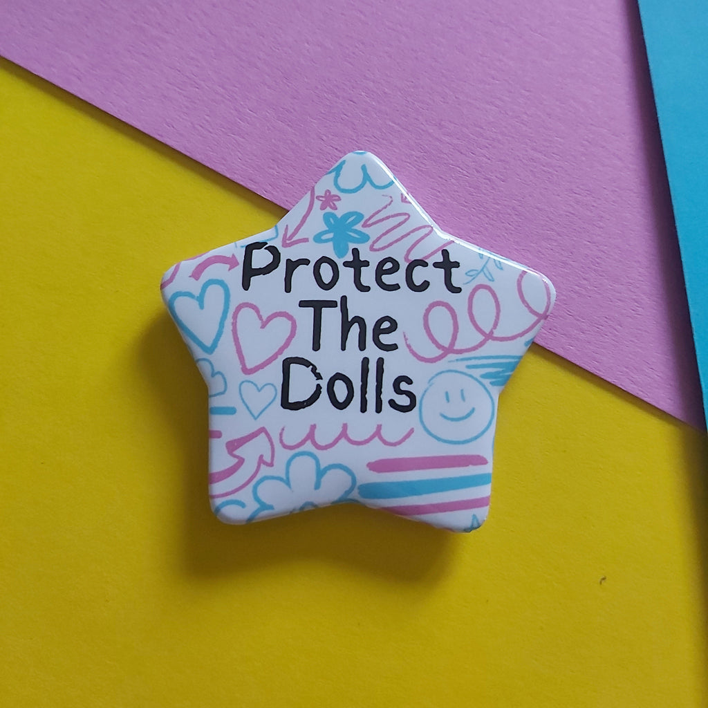 A star-shaped pin with the text 'Protect The Dolls' in black, decorated with pink and blue doodles on a white background, resting on a split yellow and purple paper backdrop.