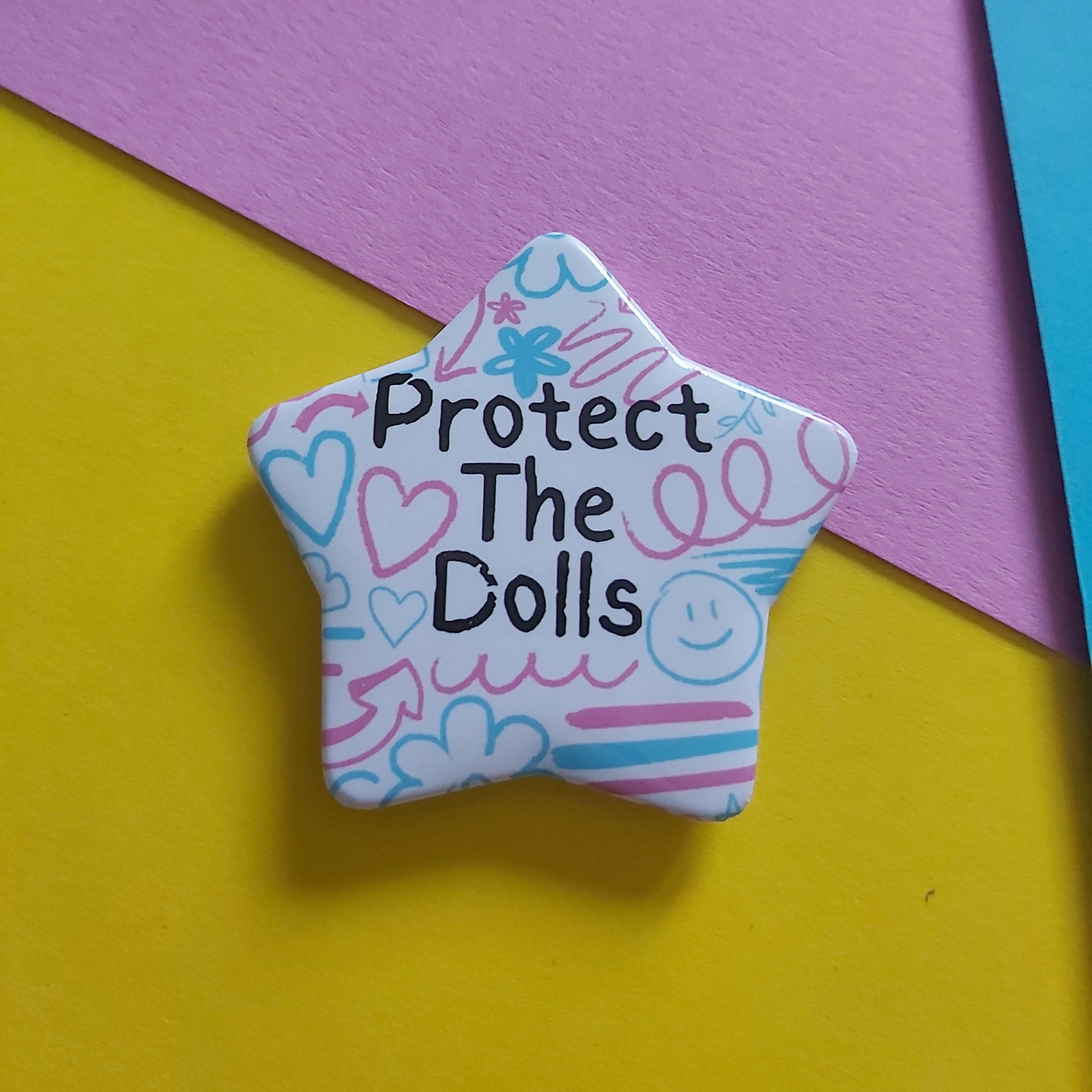 A star-shaped pin with the text 'Protect The Dolls' in black, decorated with pink and blue doodles on a white background, resting on a split yellow and purple paper backdrop.