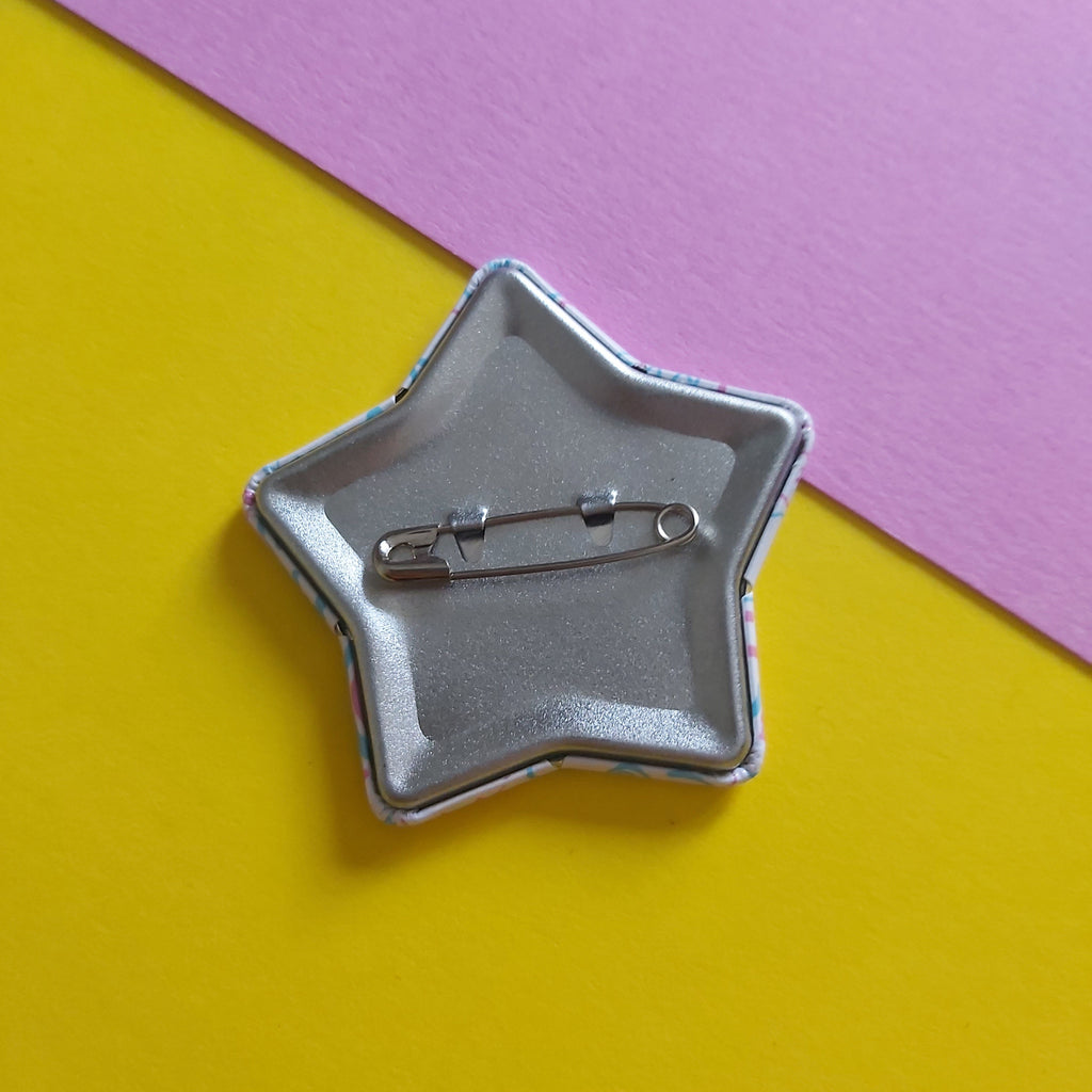 A star-shaped silver pinback button showing its safety pin back, placed on a diagonally split yellow and pink background.