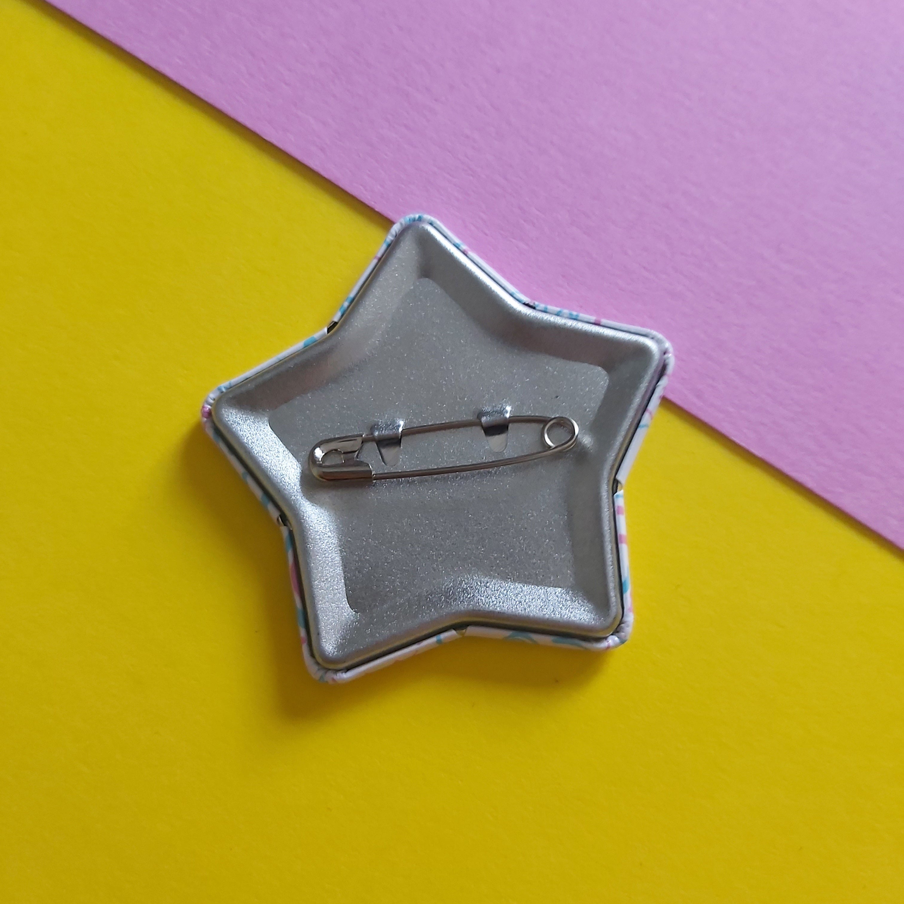 A star-shaped silver pinback button showing its safety pin back, placed on a diagonally split yellow and pink background.