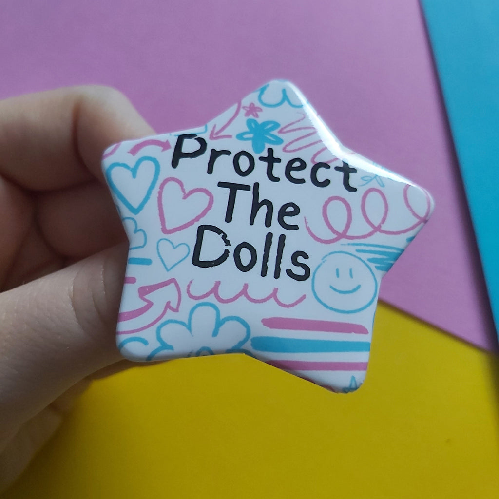 A hand holding a star-shaped pinback button with the text 'Protect The Dolls' in black, surrounded by hand-drawn pink and blue hearts, smiley faces, and doodles on a white background. The background consists of colorful paper sheets in pink, yellow, and blue.