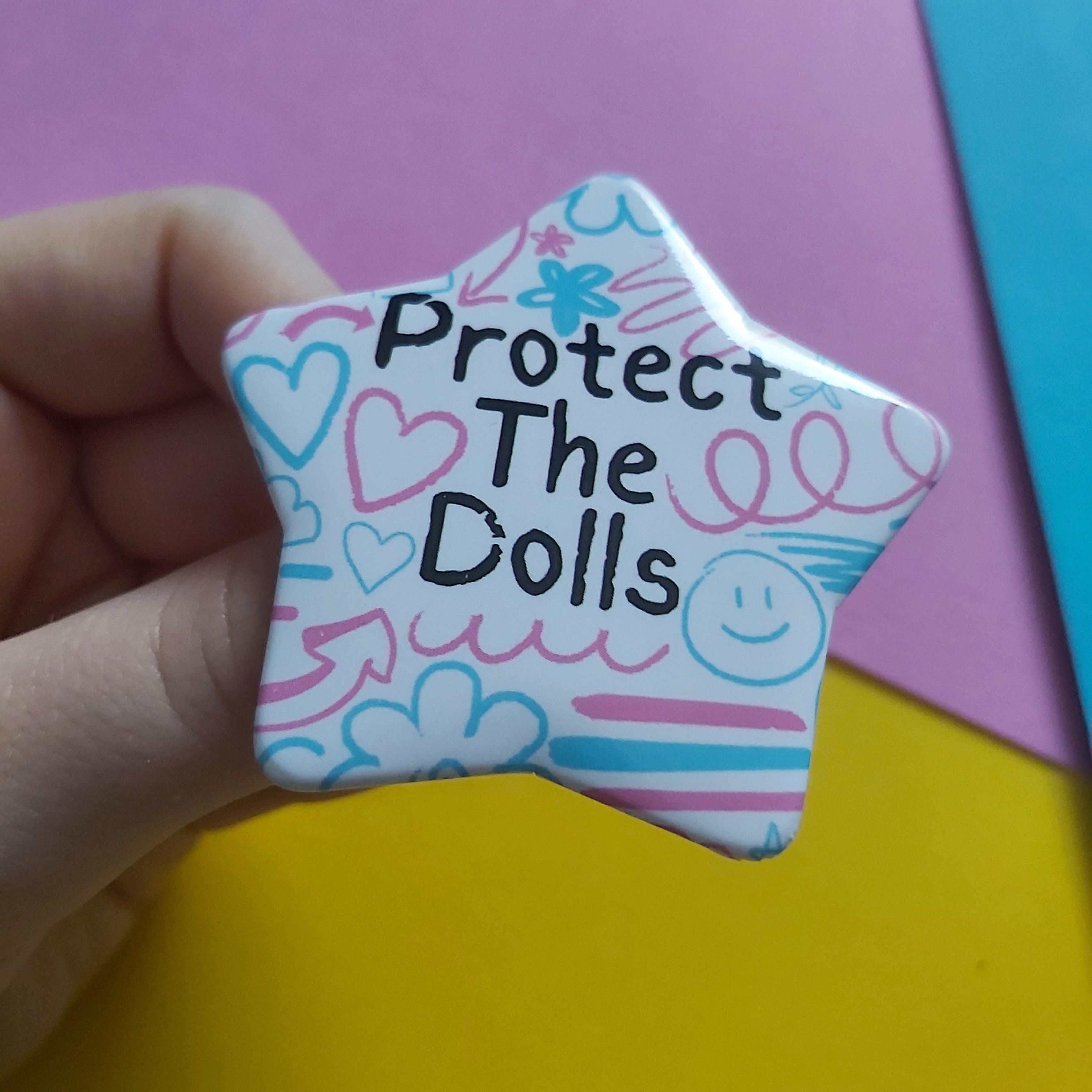 A hand holding a star-shaped pinback button with the text 'Protect The Dolls' in black, surrounded by hand-drawn pink and blue hearts, smiley faces, and doodles on a white background. The background consists of colorful paper sheets in pink, yellow, and blue.
