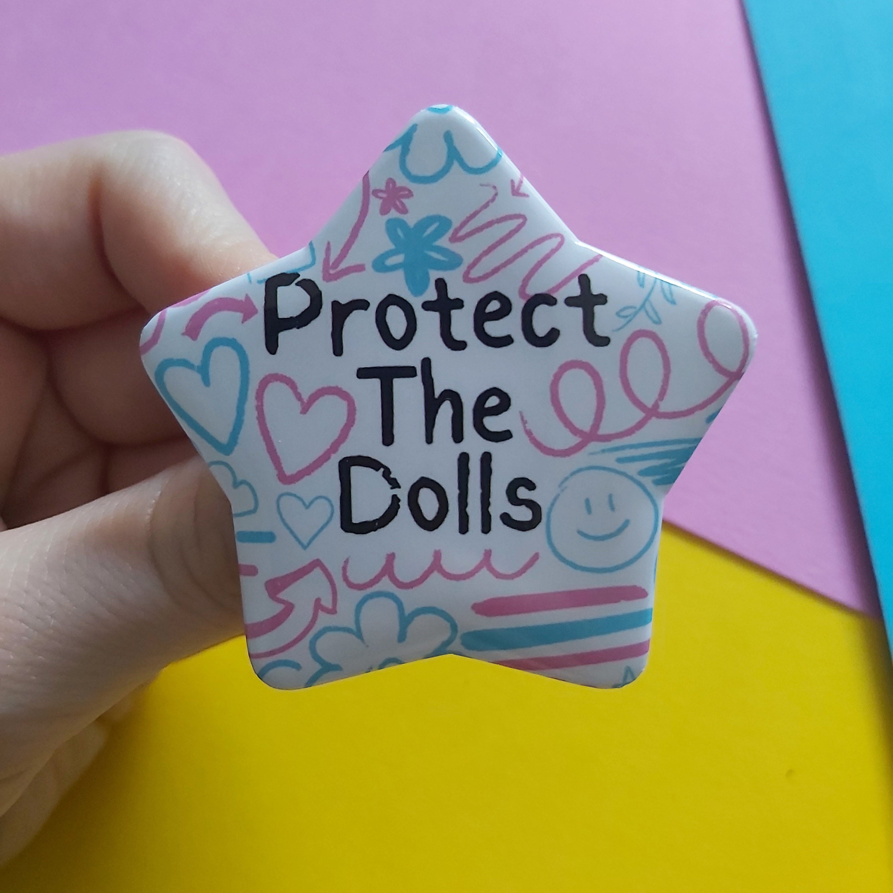 A hand holds a star-shaped pin or badge with the text 'Protect The Dolls' in black, decorated with pink and blue doodles on a white background, set against a colourful pink, yellow, and blue paper backdrop.