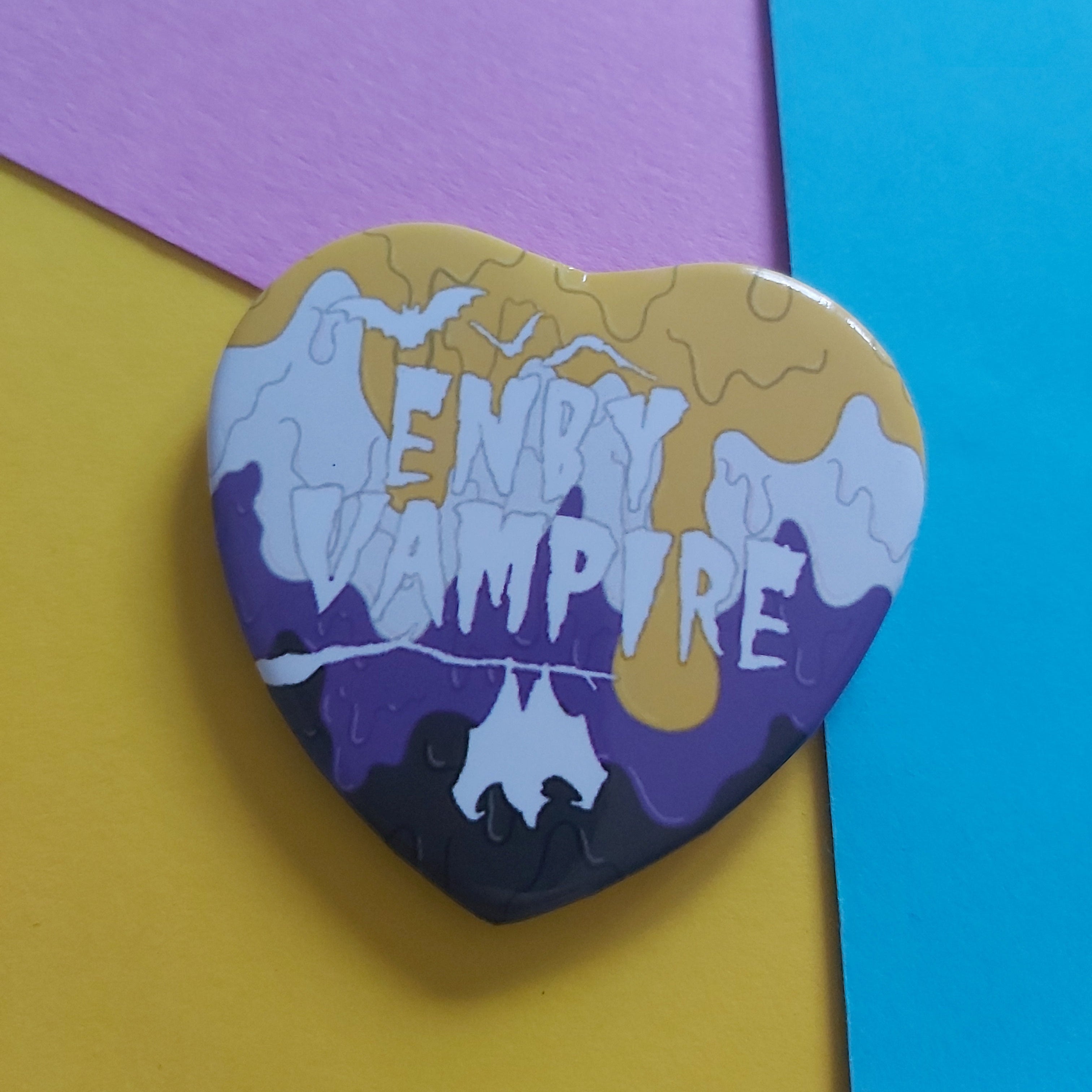A heart-shaped pin featuring a multicolour paint design with the words 'ENBY VAMPIRE' in white text, displayed on a background of yellow, purple, and blue paper.