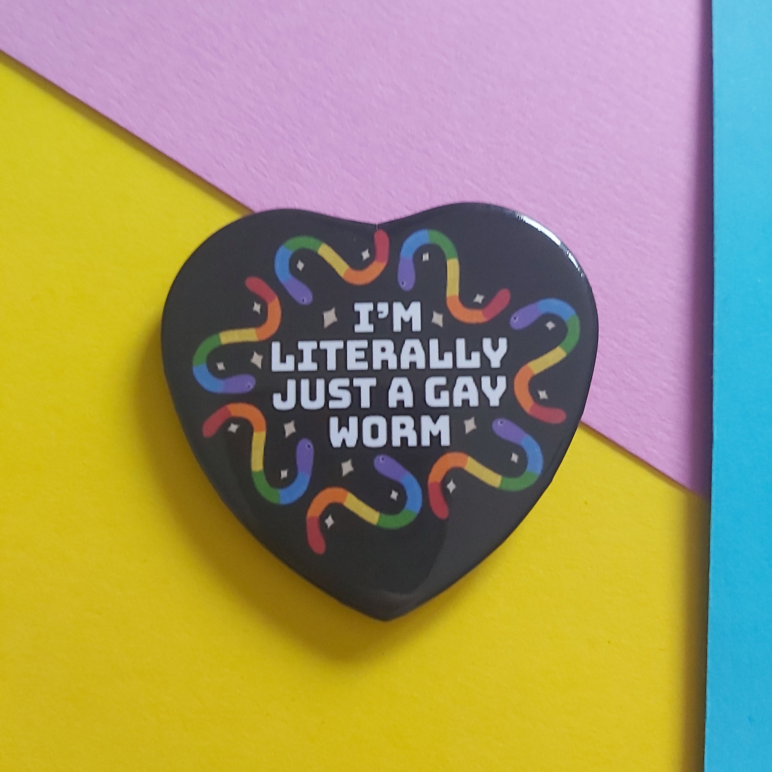 A heart-shaped black pin with the text 'I'M LITERALLY JUST A GAY WORM' in white, surrounded by rainbow-colored squiggly worms, displayed on a diagonally split yellow and pink background with a blue strip on the right.