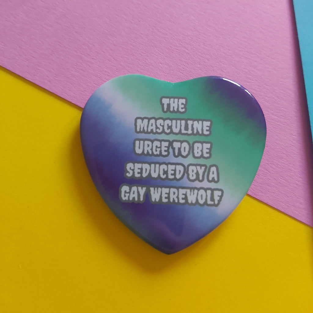 A heart-shaped pin with a blue and green gradient design featuring the text 'THE MASCULINE URGE TO BE SEDUCED BY A GAY WEREWOLF' in white. The pin is displayed on a background of pink, yellow, and light blue paper.