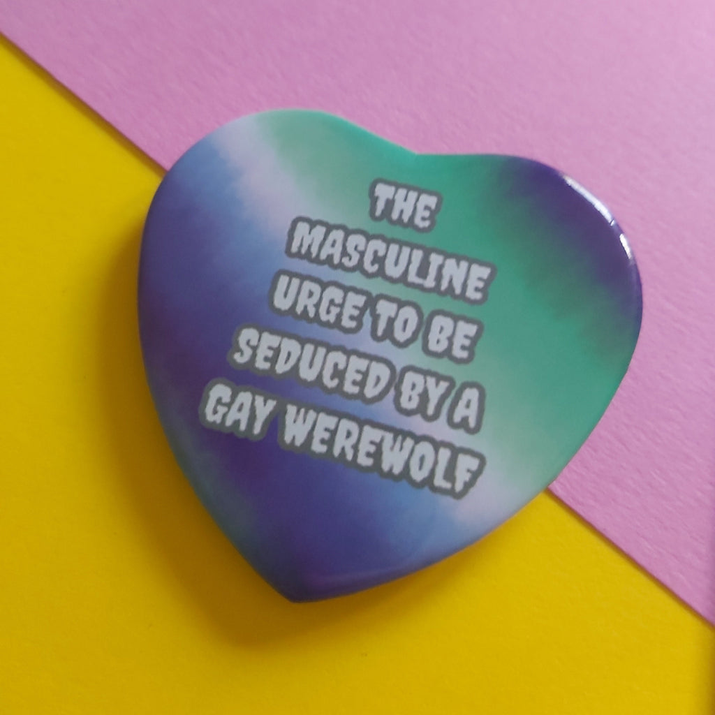 A heart-shaped pin badge featuring a blue and green gradient design with the text 'THE MASCULINE URGE TO BE SEDUCED BY A GAY WEREWOLF' in white with a black outline, set against a split pink and yellow background.
